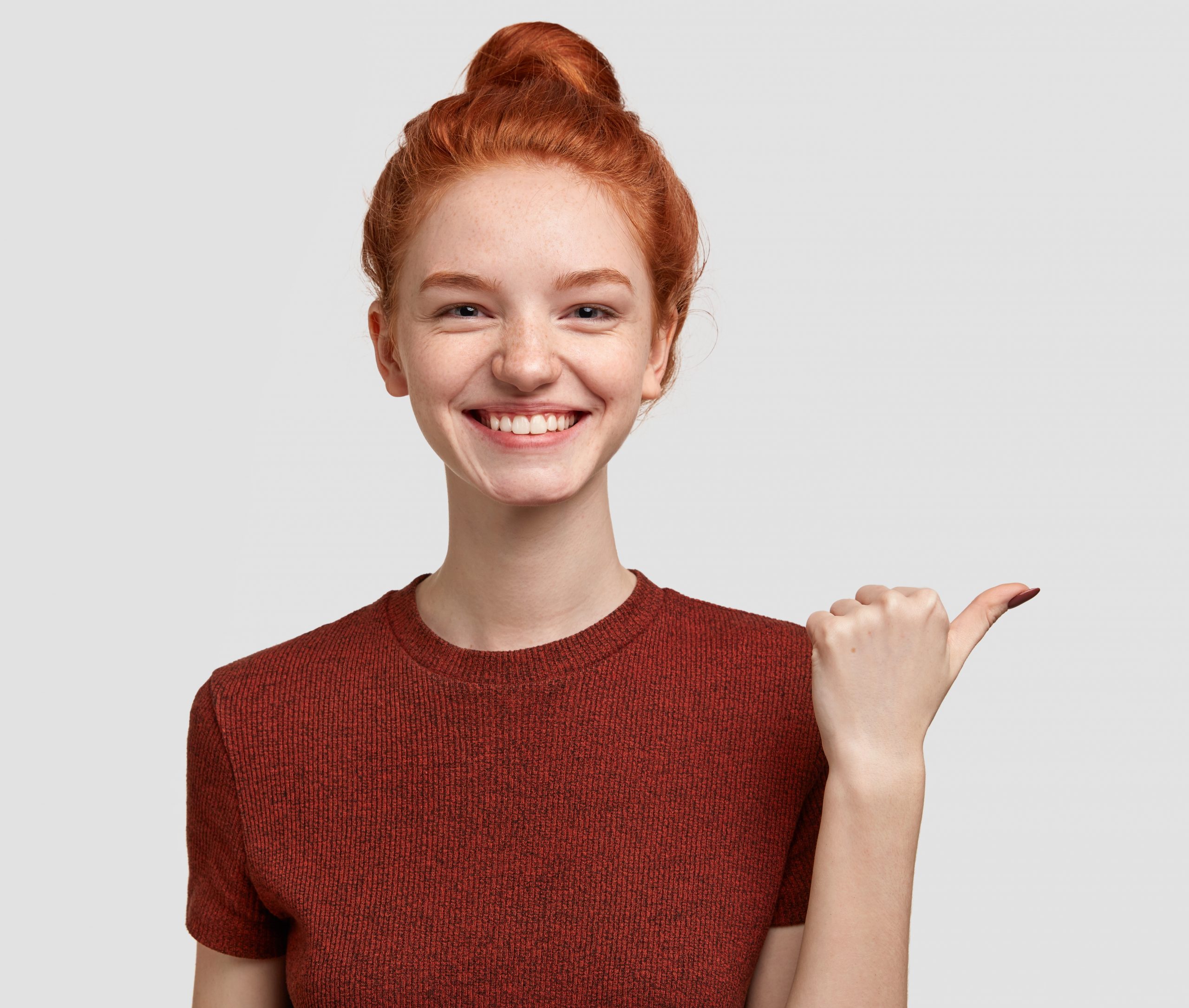 Advertisement concept. Cheerful foxy girl with tender toothy smile, freckled skin indicates with thumb aside, shows something over blank space, dressed in casual outfit, isolated on white background.